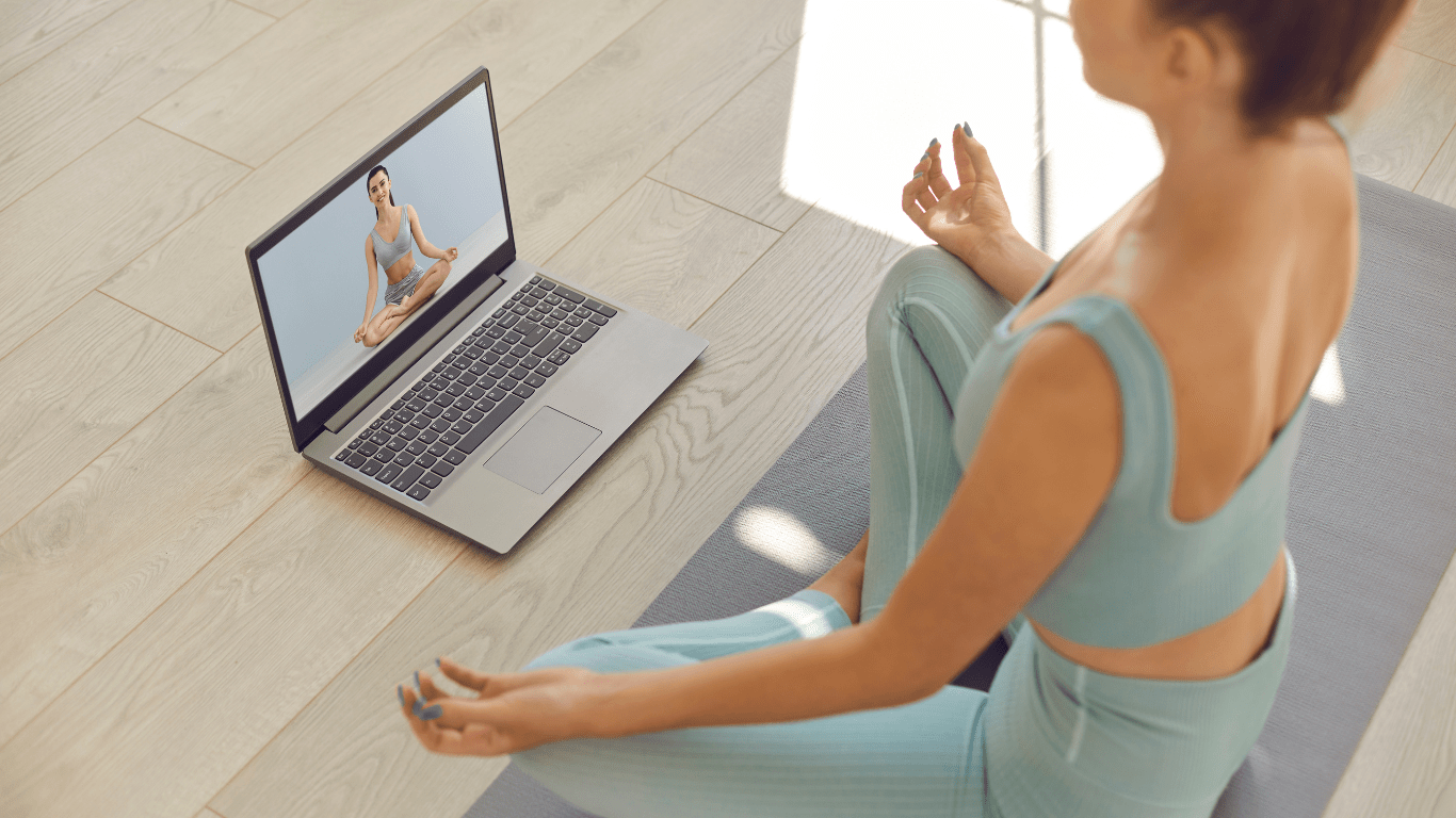 A person is meditating on a yoga mat in front of a laptop